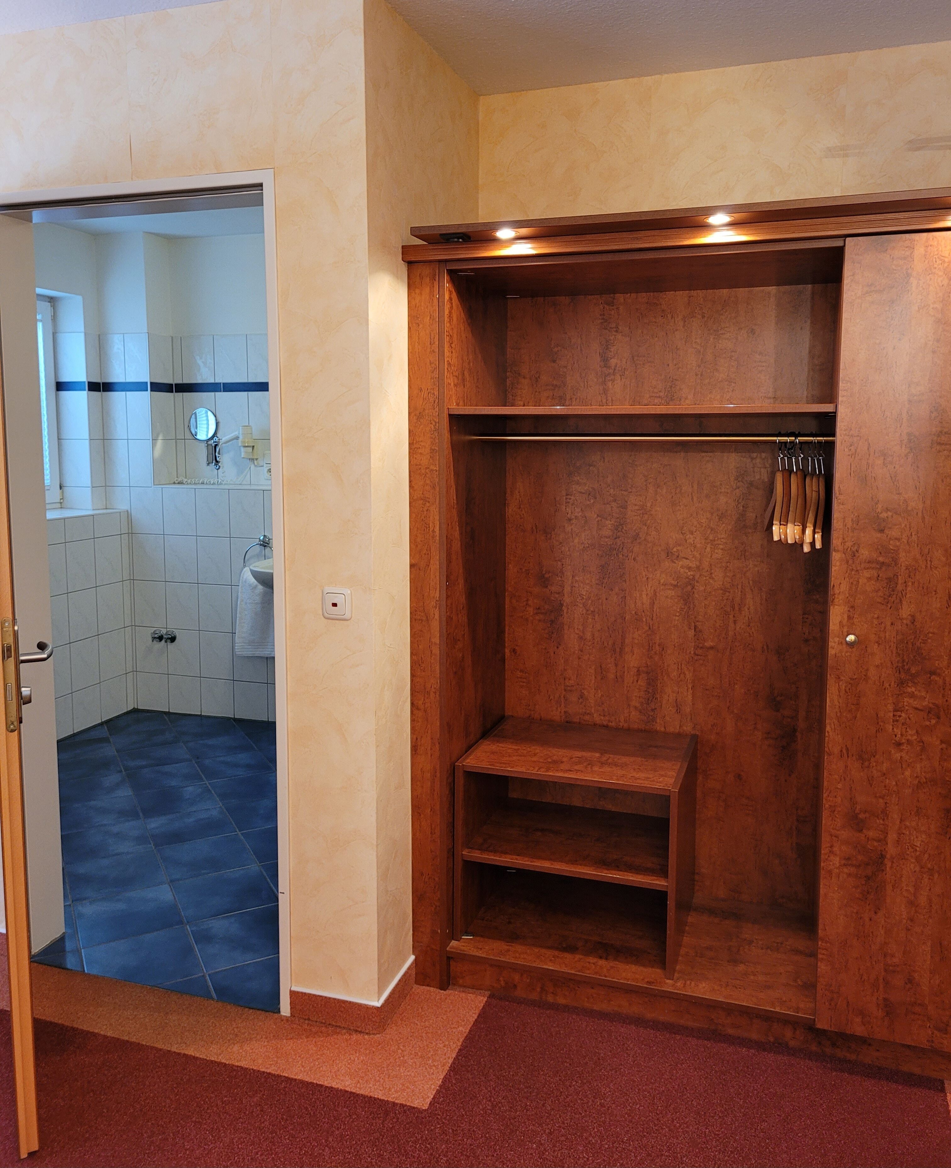 Ein Holzkleiderschrank mit Spiegel und Haken, einem kleinen Ablagefach und einer Tür zu einem Badezimmer mit Fliesenboden.