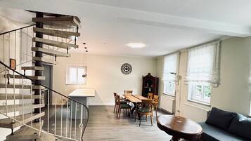 Een ruime woonkamer met een houten trap, een eettafel met stoelen, een bank en een ronde salontafel.