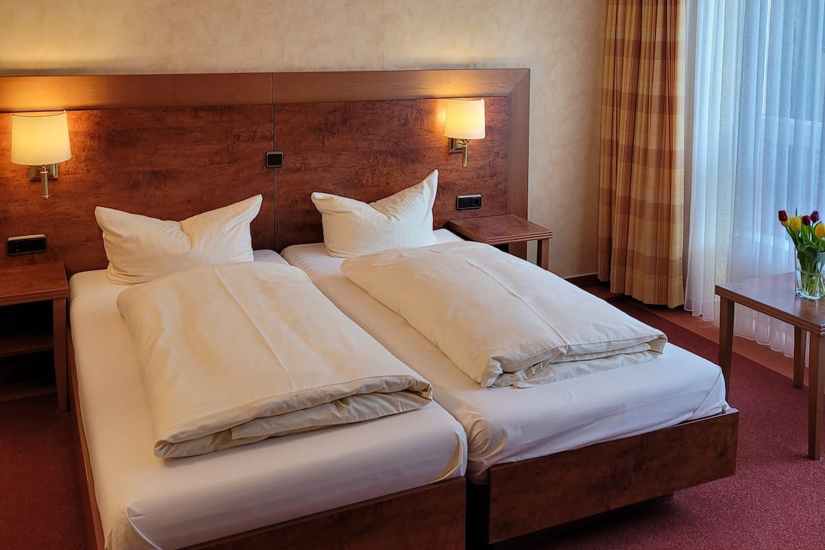 A double bed with two pillows, a wooden headboard, two bedside tables with lamps, a small table with a vase of flowers, and a window with curtains.
