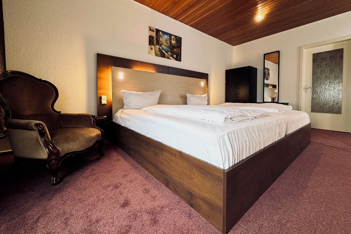 A hotel room with a large bed, a wooden headboard, a chair, and a painting on the wall.