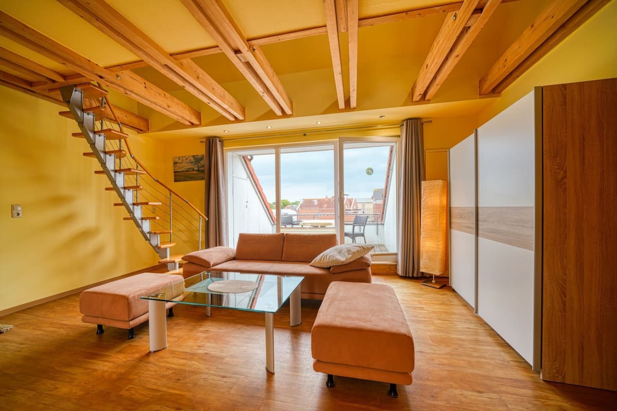 A spacious living room with a glass coffee table, a sofa, and a cushioned bench. A wooden staircase with orange handrails leads to a balcony with a view of buildings.