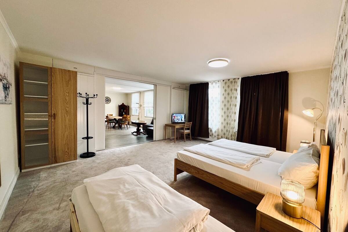 A hotel room with two beds, a desk, a TV, and a dining area visible through an open door.