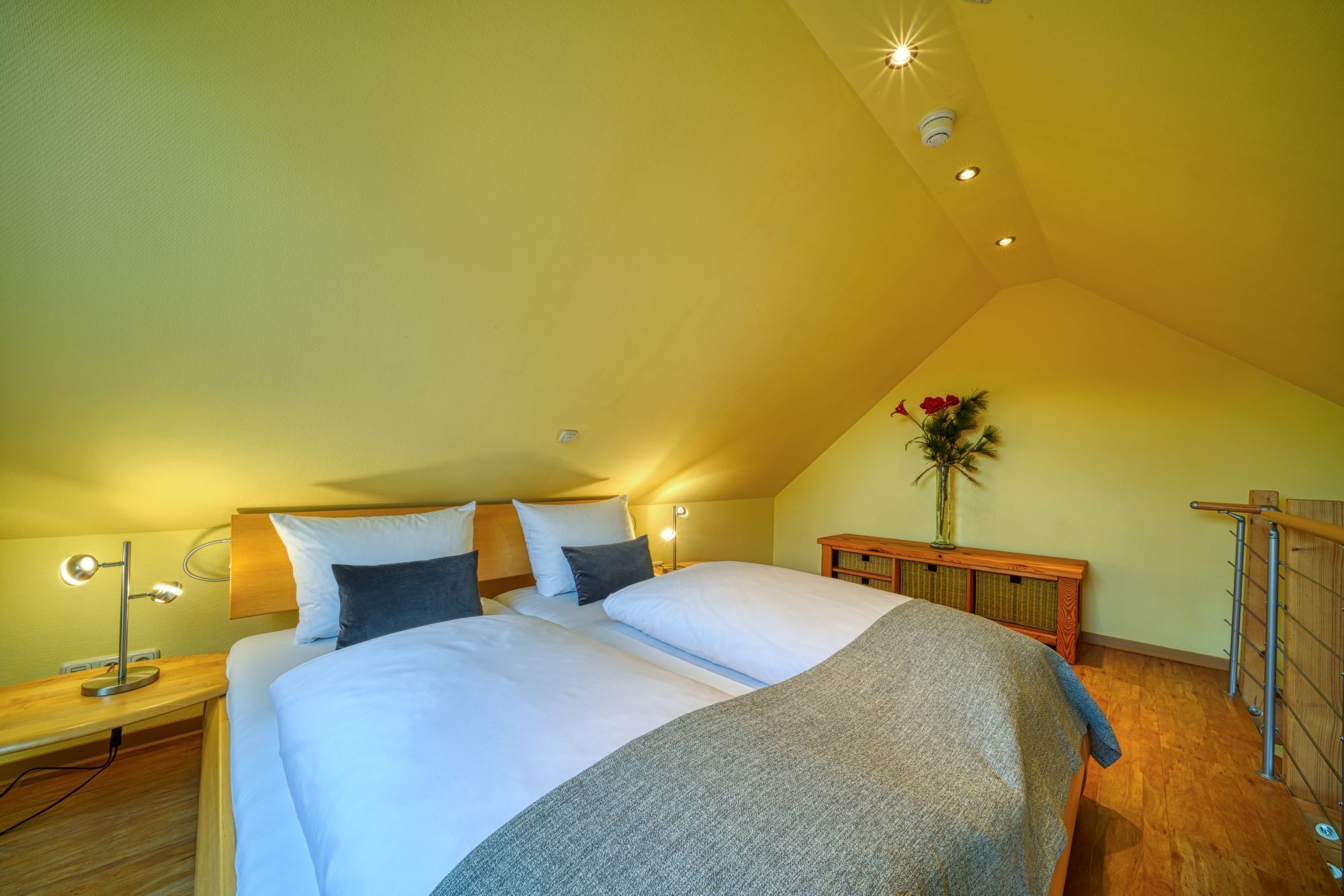 A bedroom with a large bed, a bedside table, a lamp, a wooden headboard, a vase with flowers, and a wooden railing.