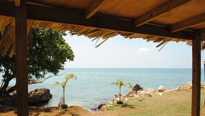 Una estructura de madera con techo de paja que domina una playa de aguas azules y algunas palmeras.