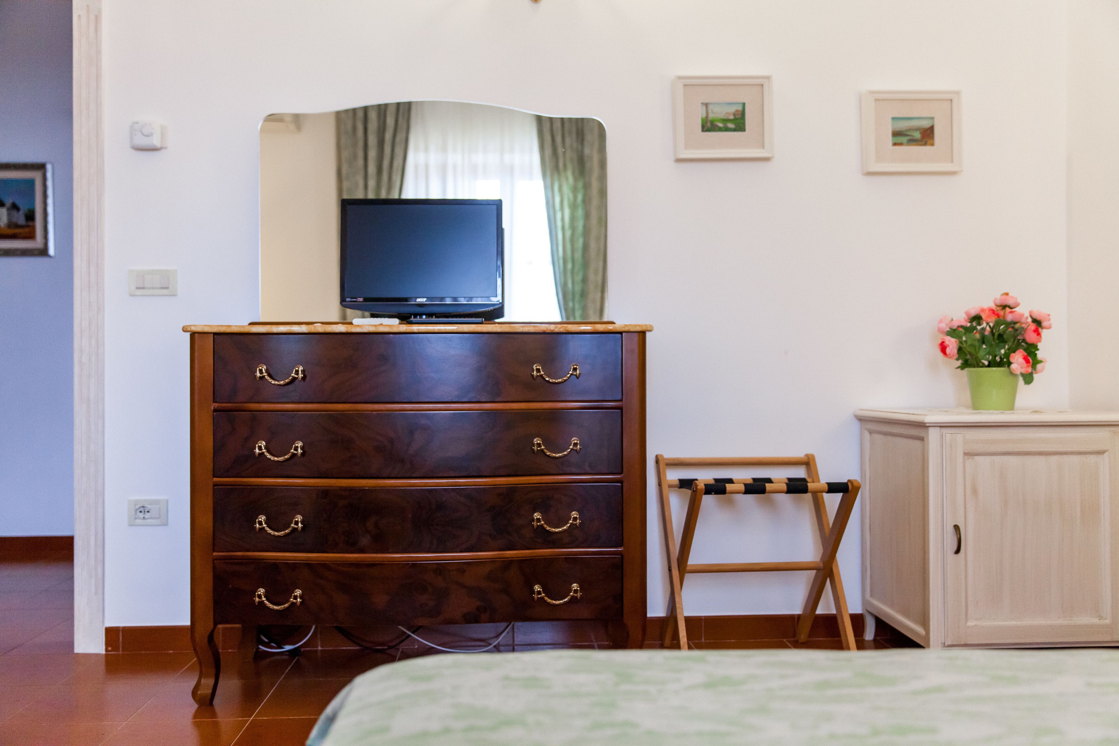 A room with a wooden dresser, a television on top, a small table with a vase of flowers, and a bed with a green and white patterned bedspread.