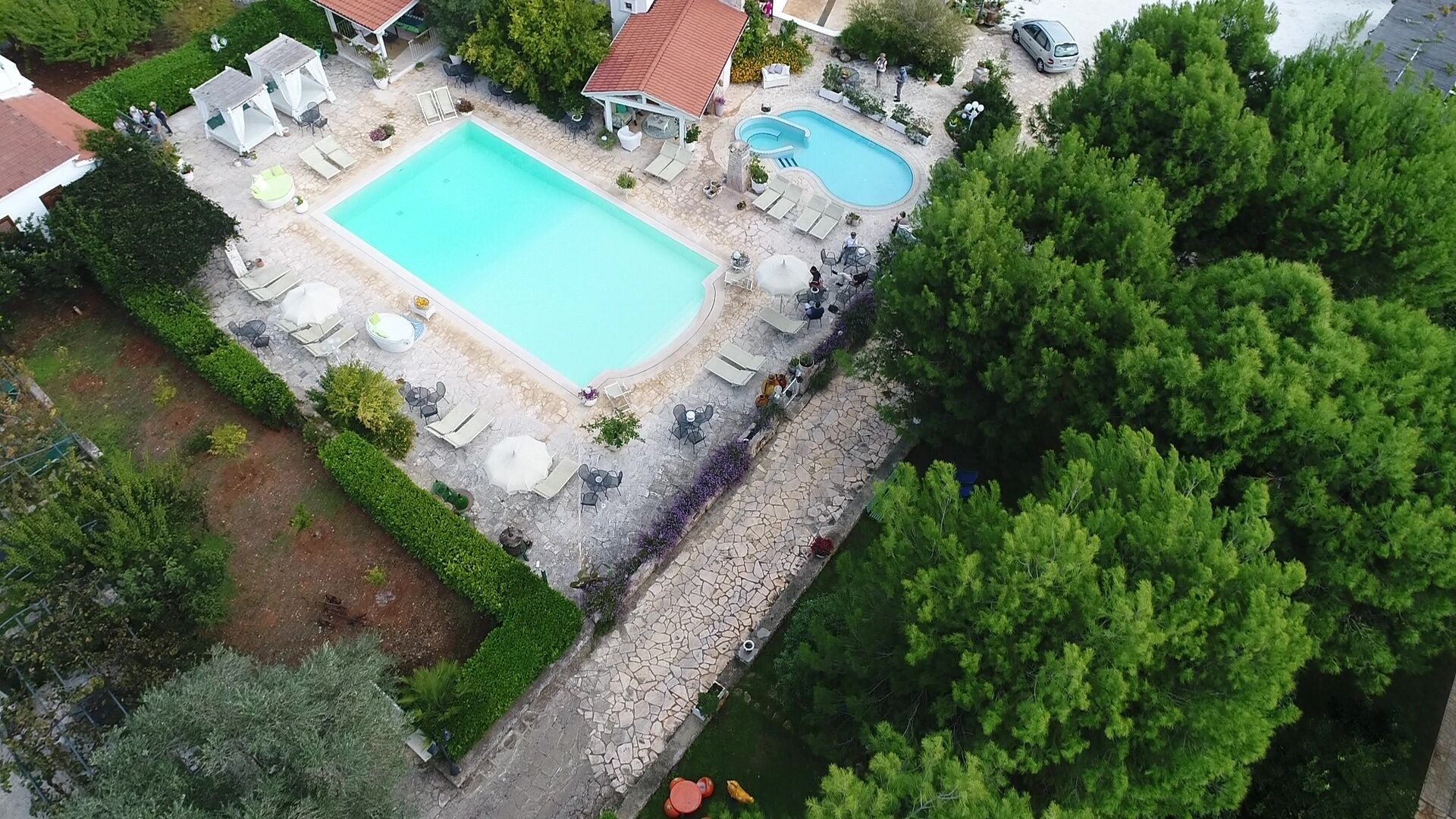 An aerial view of a resort with a large rectangular swimming pool, a smaller circular pool, and a covered seating area with umbrellas.