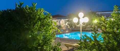 Una piscina rodeada de exuberante vegetación, con un edificio al fondo e iluminación exterior.