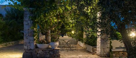 Eine Terrasse im Freien mit Steinsäulen, einem Tisch und Stühlen, umgeben von Grünpflanzen und Steinmauern.