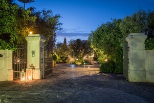 A paved courtyard with a gated entrance, surrounded by greenery and illuminated by lanterns.
