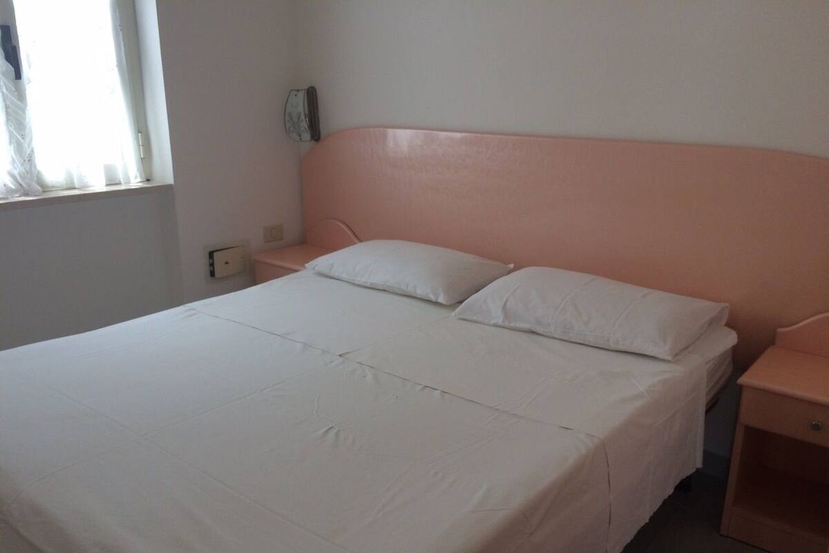 Un lit bien fait, avec du linge de lit blanc, dans une chambre sobre agrémentée d’une tête de lit rose.