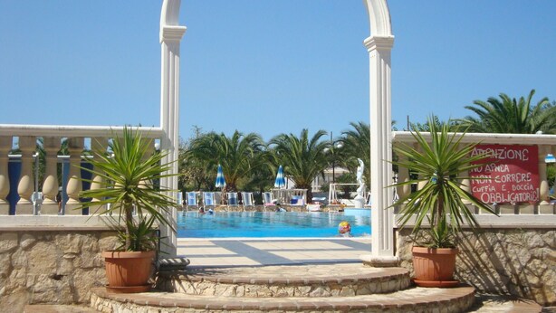 A pool area with a white archway, palm trees, and a sign with text. - Villaggio Piano Grande (Vieste)