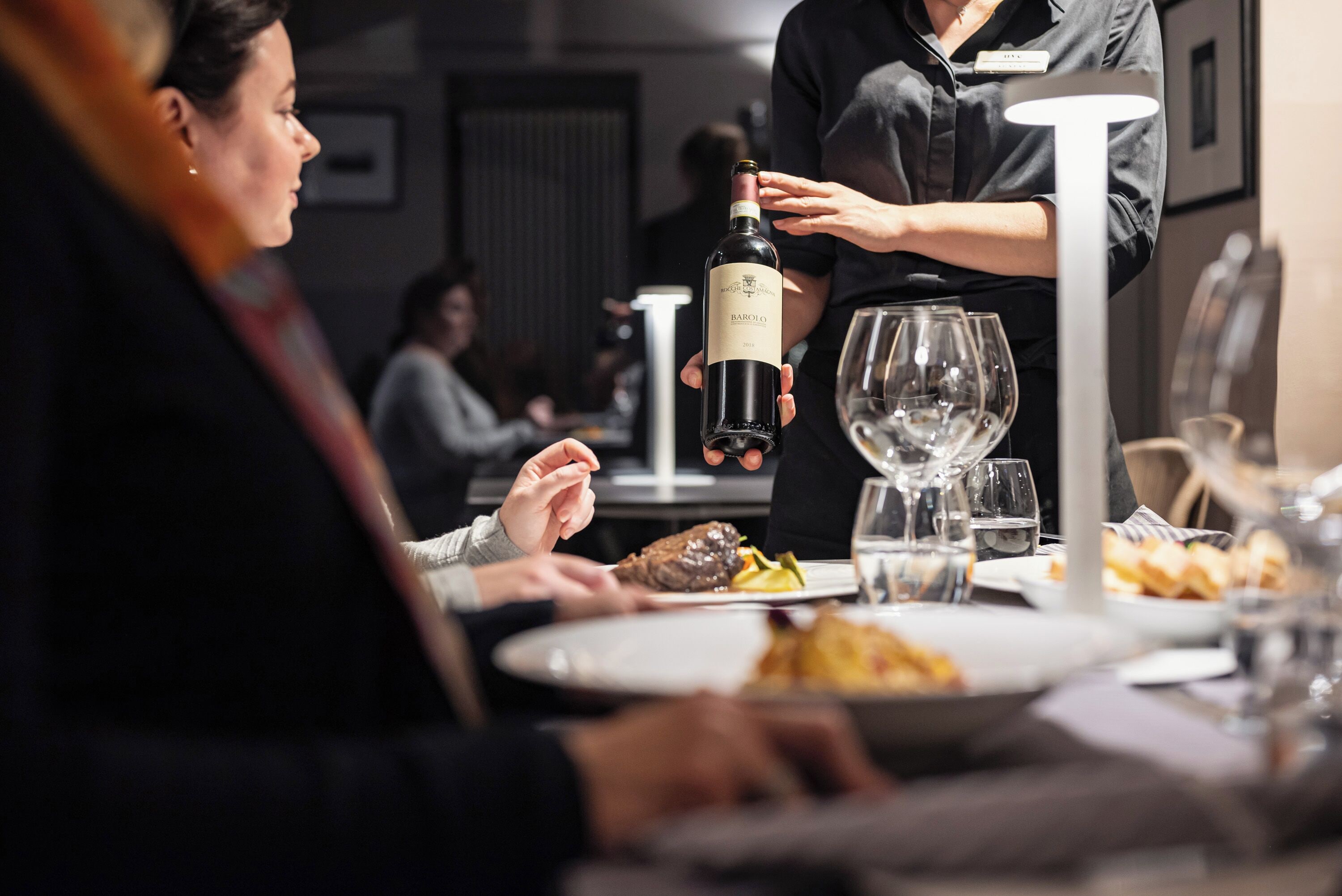 A dining table with plates of food, a bottle of wine, and a waiter serving.