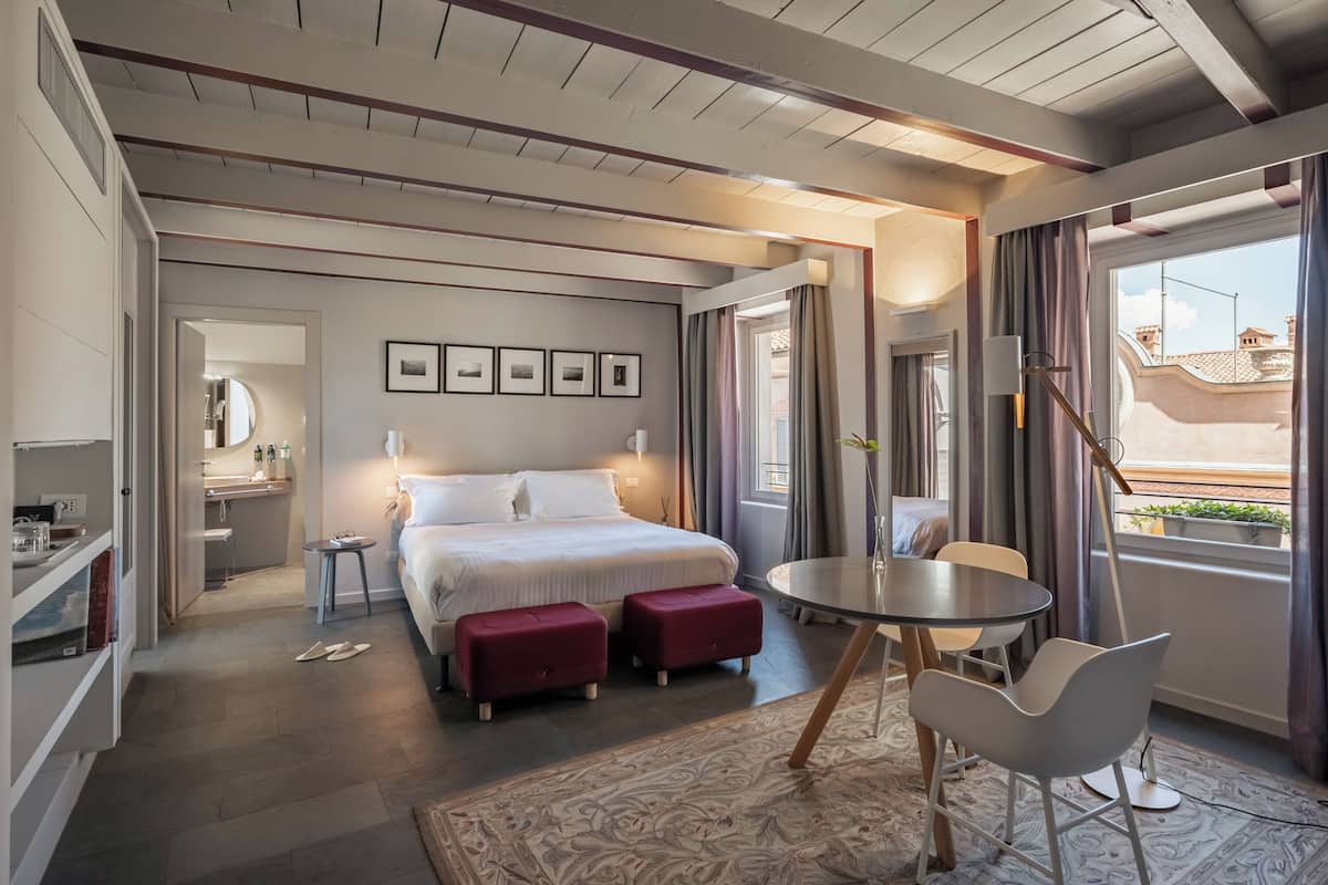 Une chambre d’hôtel moderne avec un grand lit, une petite table et une chaise. La salle de bain est équipée d’un miroir et d’un lavabo. La chambre offre une vue sur les bâtiments situés à l’extérieur, visibles par la fenêtre.