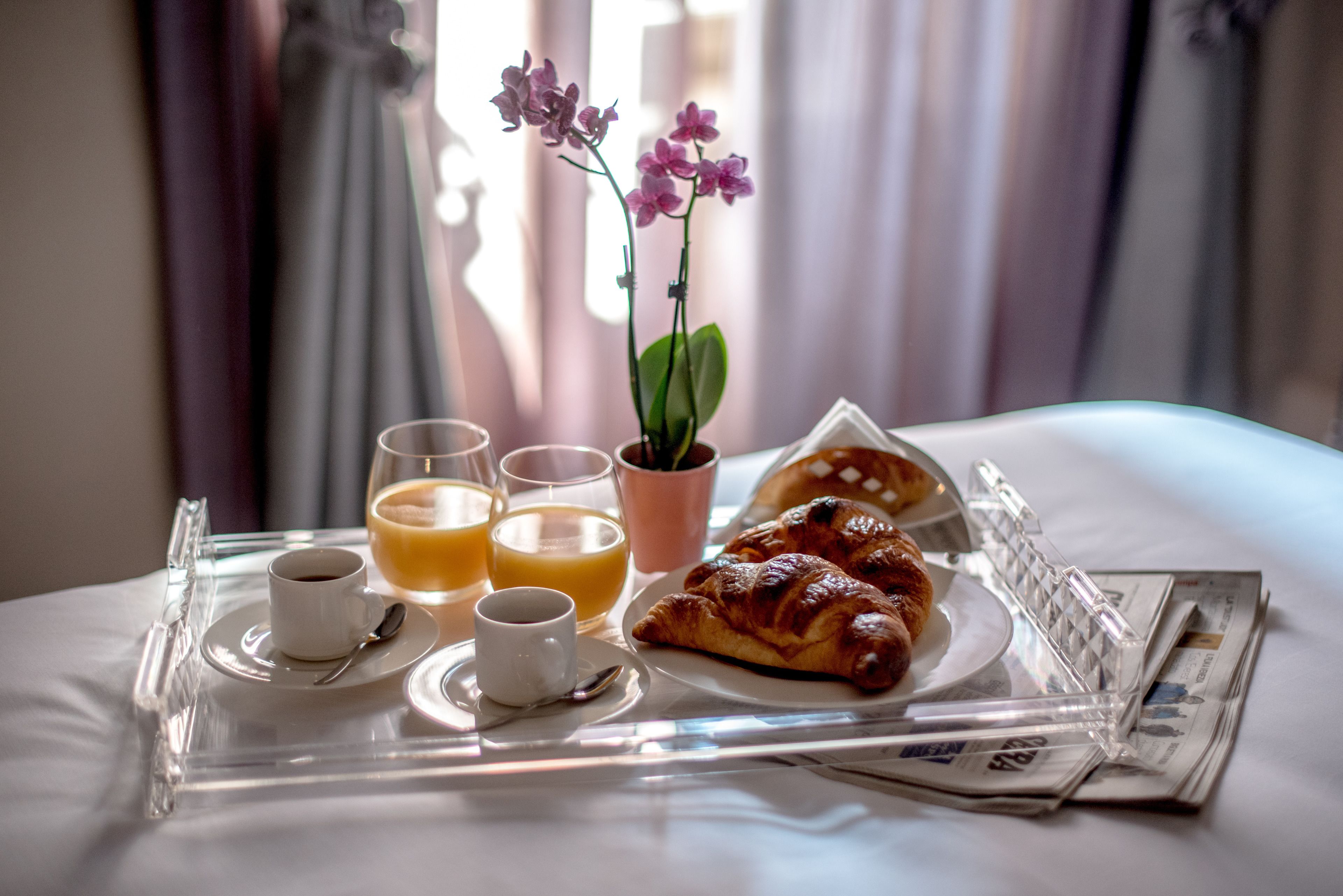 A breakfast tray with croissants, coffee, orange juice, and a potted orchid on a bed.