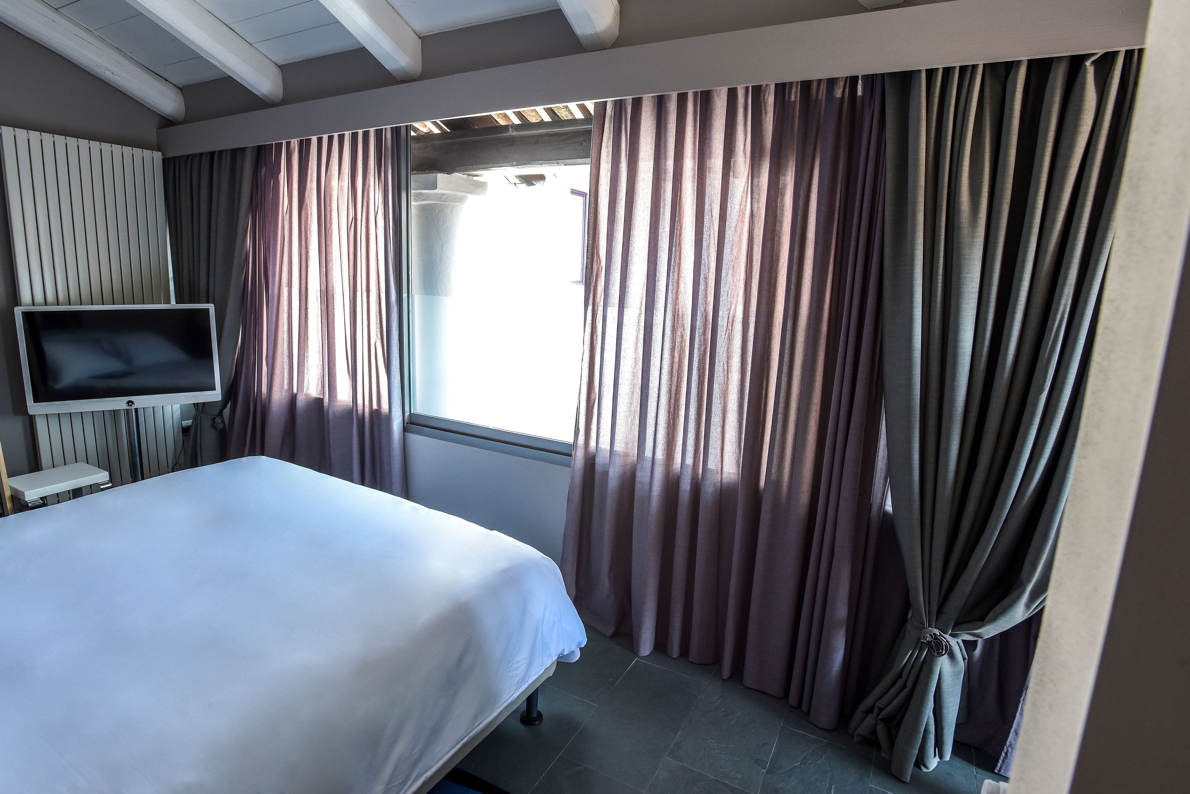 A hotel room with a bed, a TV mounted on the wall, and a window with curtains.