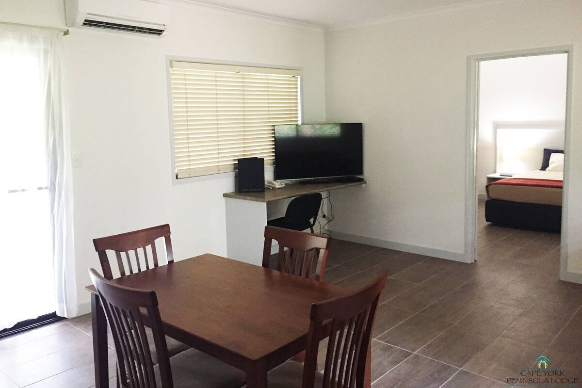 A room with a dining table and chairs, a television on a stand, and a bed in the adjacent room.