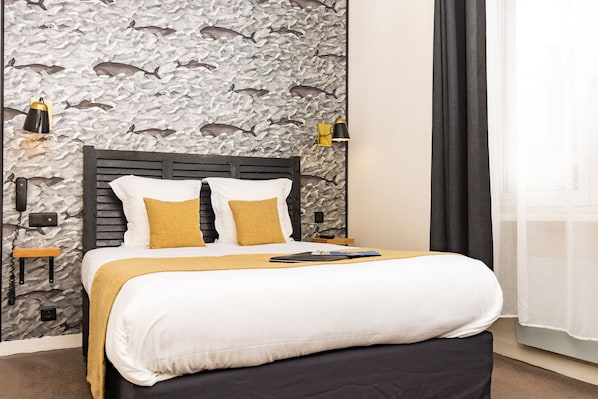 A neatly made bed with white and mustard bedding, a wooden headboard, and a wall with a fish pattern. - Hôtel Les Charmettes (Saint-Malo)