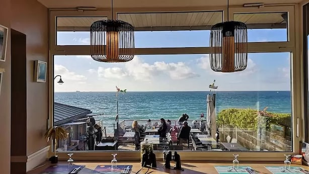 Ein Restaurant mit großen Fenstern, die auf das Meer blicken, mit hängenden Lampen und Außenbereich.