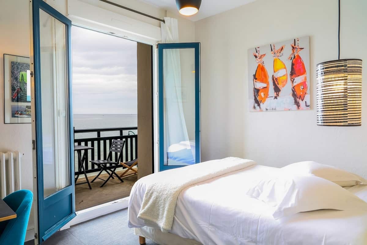 Een hotelkamer met een bed, een balkon met uitzicht op zee, een schilderij aan de muur en een lamp.