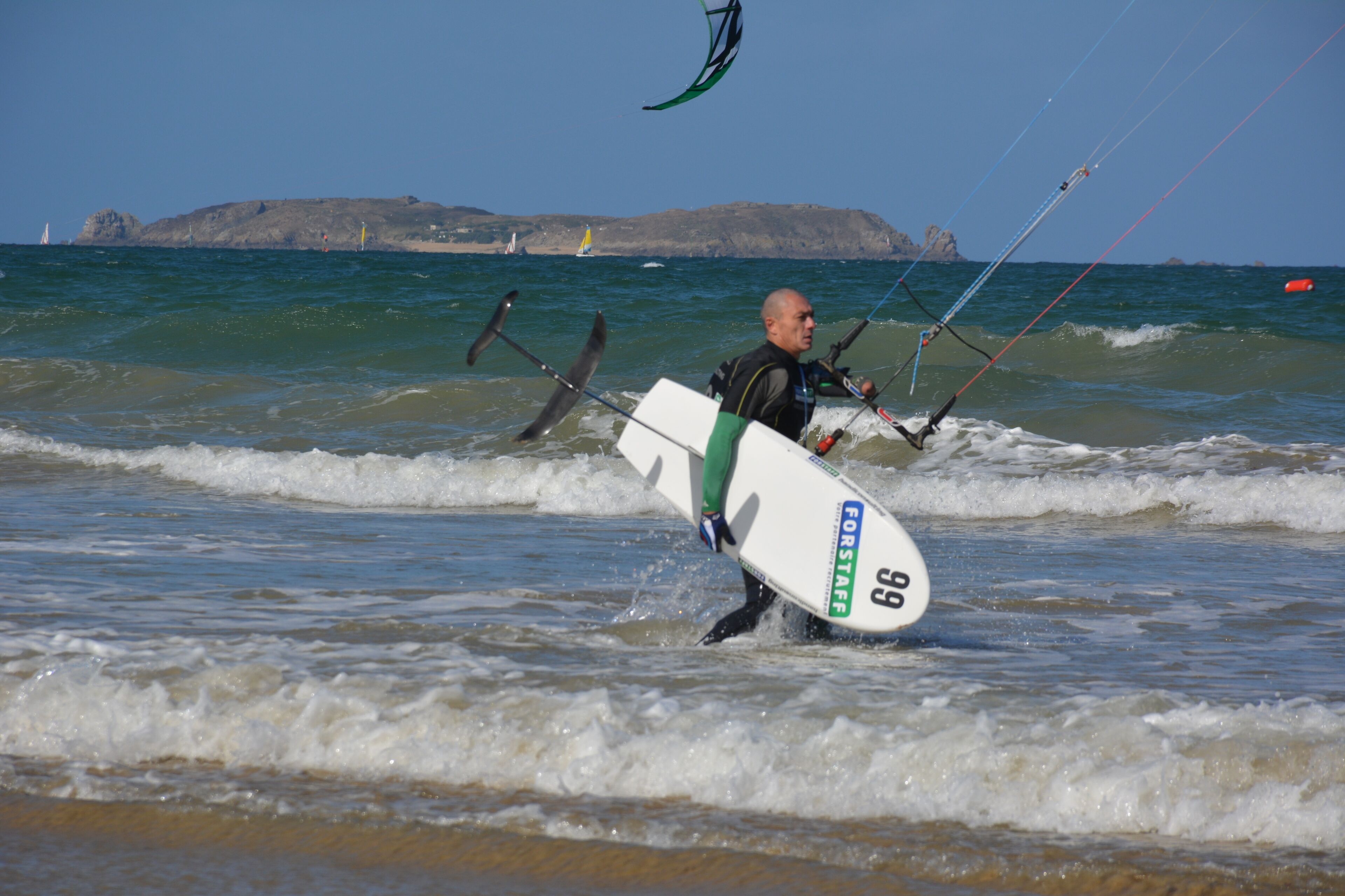 Un hombre lleva una tabla de surf con el número 99 y la palabra "FORSTAFF" caminando por el agua.