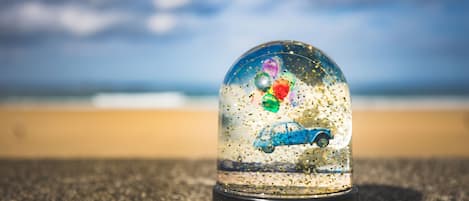 Un globo de nieve con un coche y objetos de colores en su interior, sobre una superficie rugosa, con una playa y el océano al fondo.