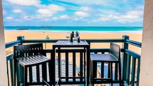 Ein Balkon mit zwei Holzstühlen und einem kleinen Tisch, der auf einen Strand mit klarem, blauem Wasser und einem Sandufer Blick bietet.