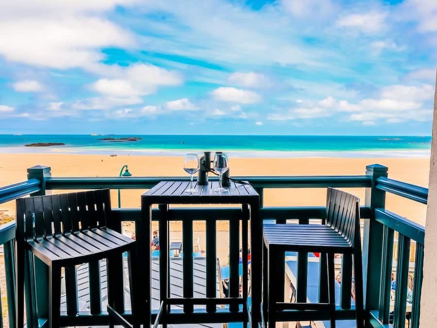 A balcony with two wooden chairs and a small table, overlooking a beach with clear blue water and a sandy shore.