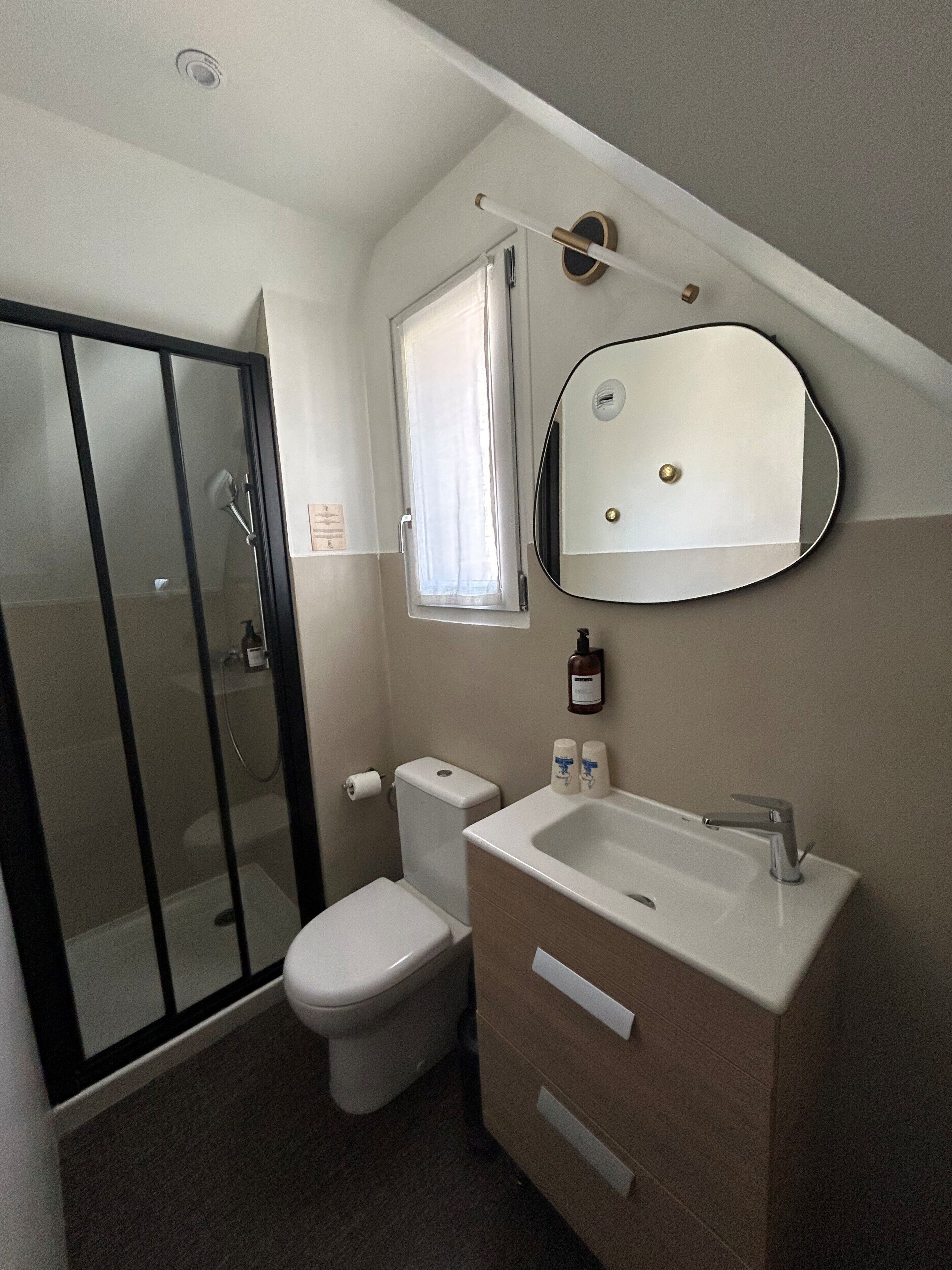 Une salle de bain avec une douche à parois de verre, des toilettes, un lavabo avec un miroir et une fenêtre.
