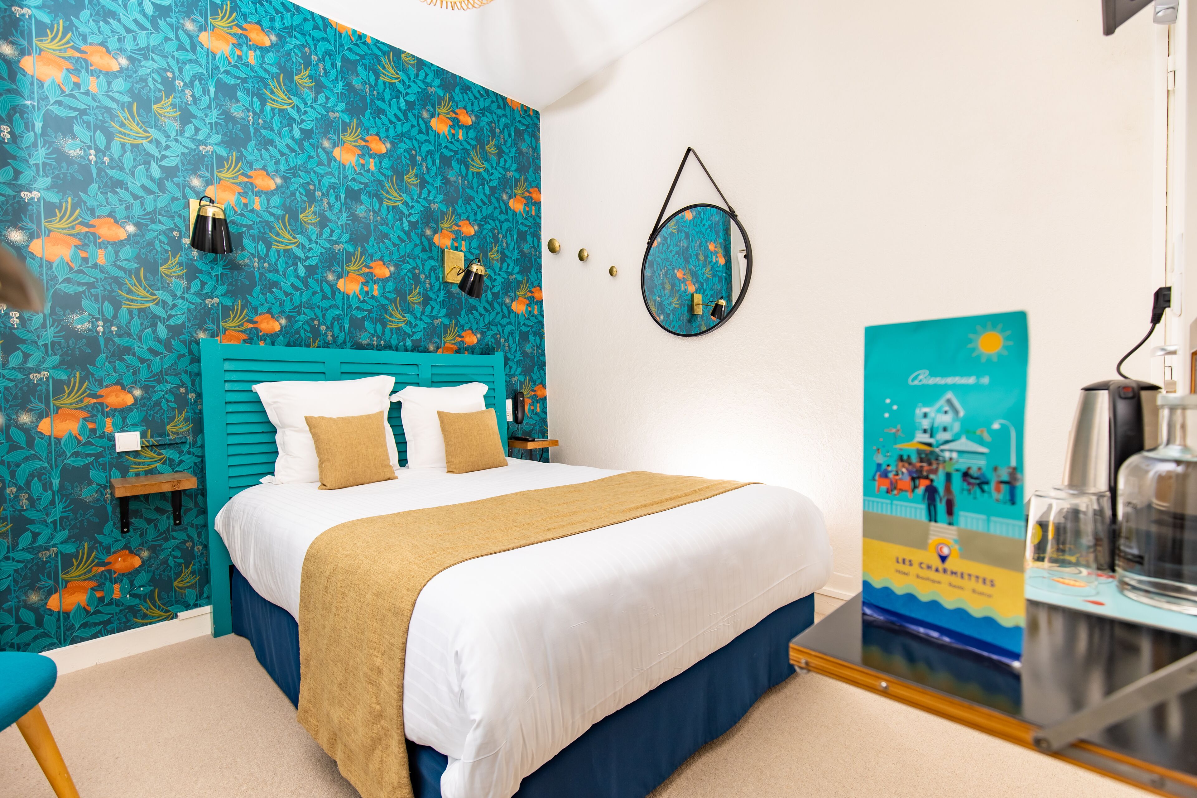 Una habitación de hotel con una cama, cabecera azul, papel tapiz floral color turquesa, un espejo redondo, un cartel y una cafetera.