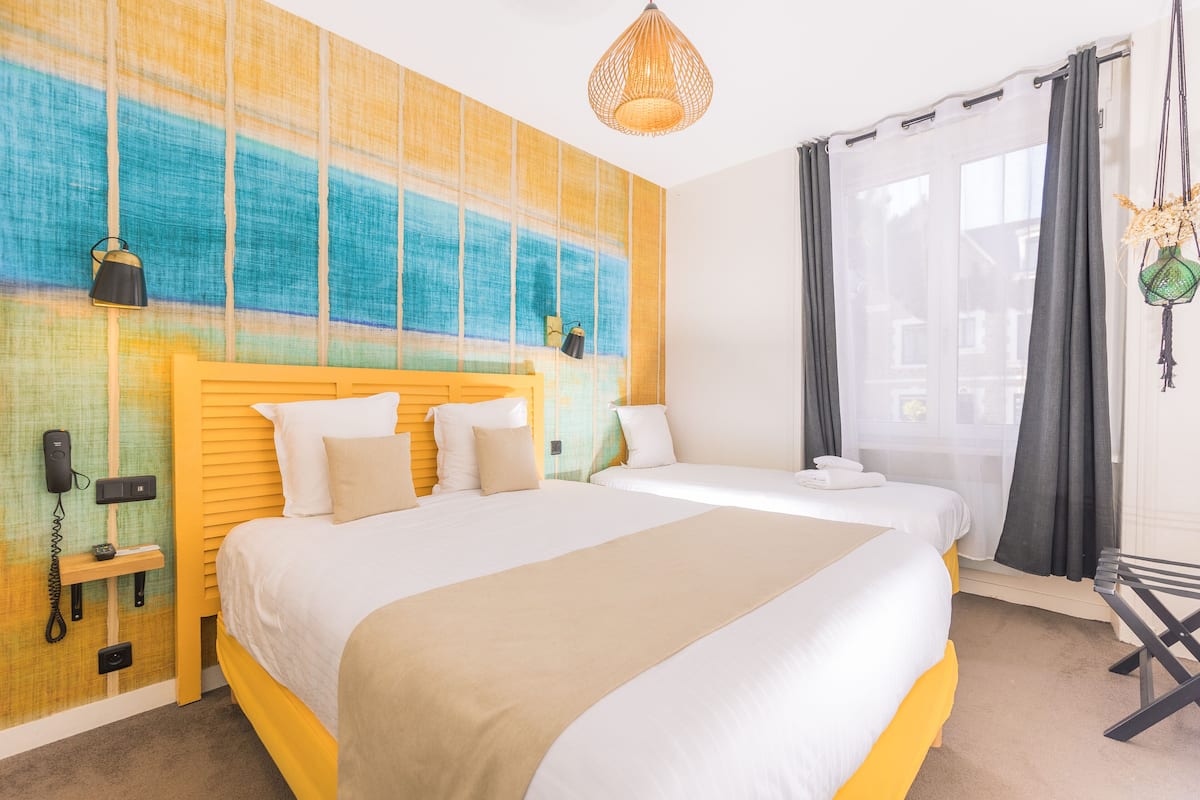 Een hotelkamer met twee bedden, een groot abstract schilderij, een hanglamp en een raam met gordijnen.