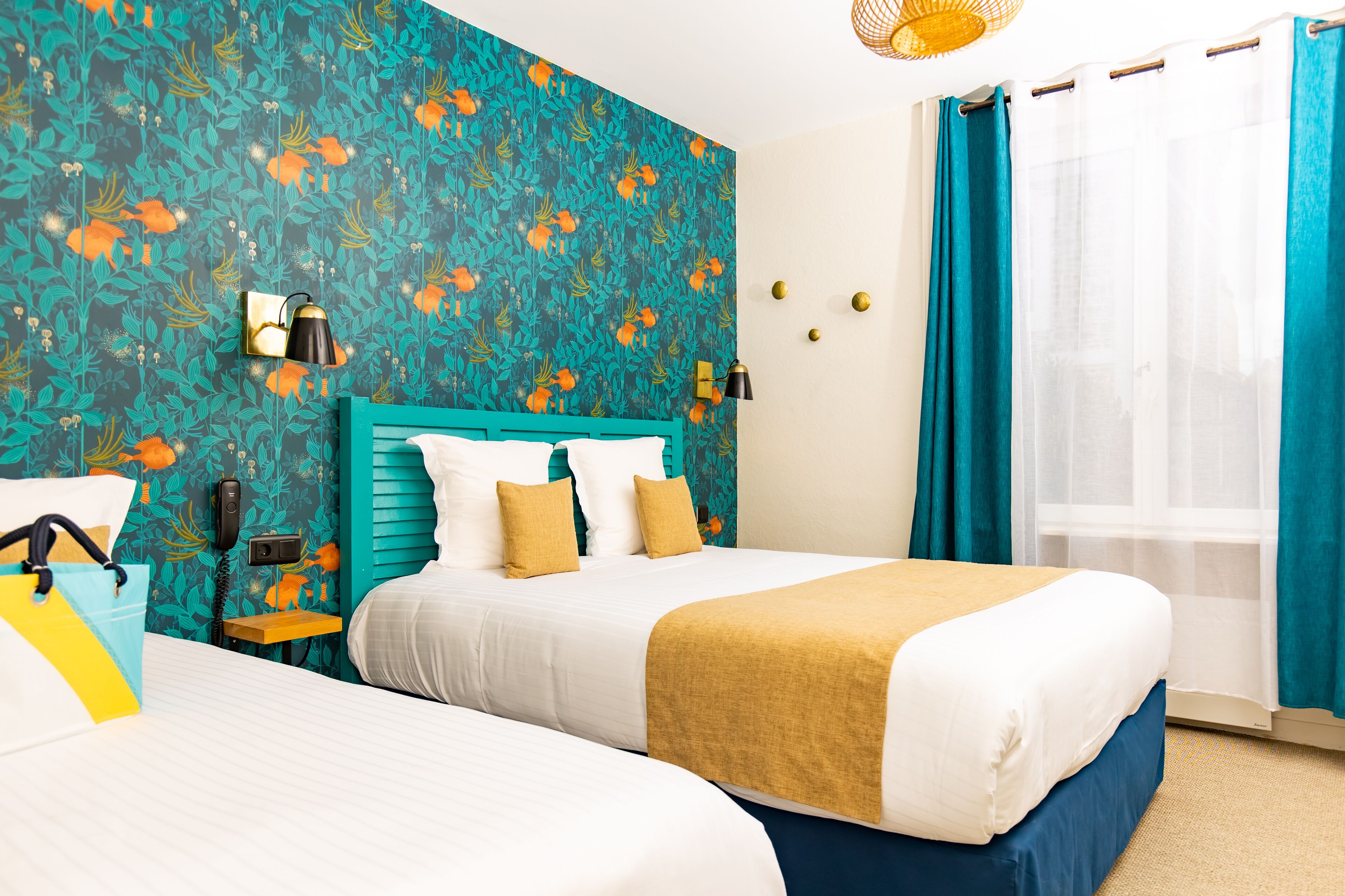 Habitación de hotel con dos camas, papel tapiz floral color turquesa, cabecera de madera y ventana con cortinas turquesas.