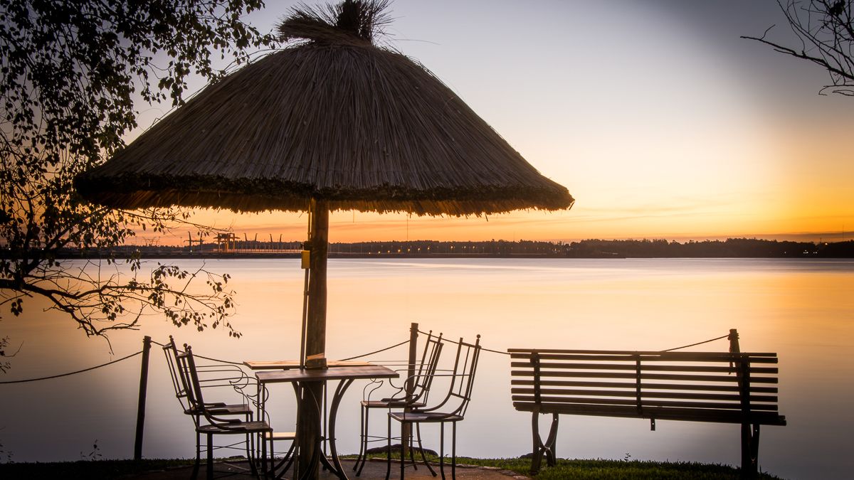 Una mesa y sillas bajo un toldo de paja junto a un lago tranquilo al atardecer.