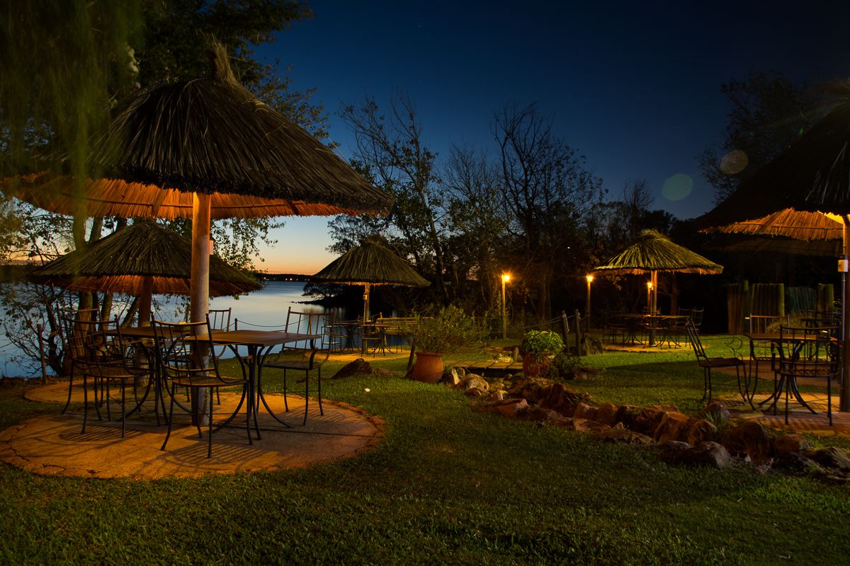 An outdoor dining area with thatched umbrellas and tables by a body of water at dusk.