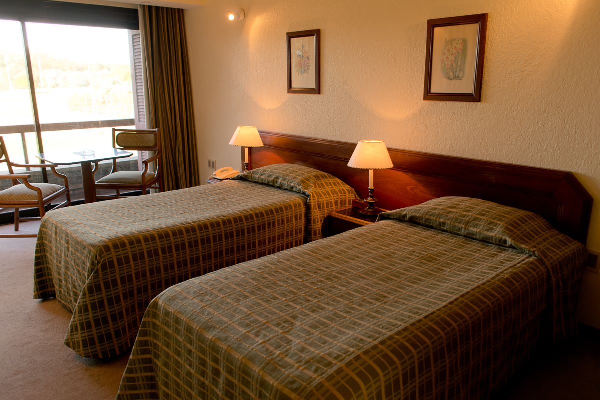 Ein Hotelzimmer mit zwei Betten, einem Holzkopfstück, zwei Nachttischlampen, einem Telefon und einem Fenster mit Vorhängen.