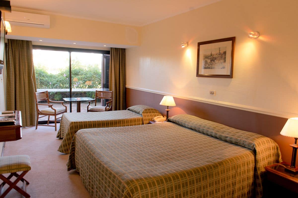 A hotel room with two beds, a small table, chairs, and a balcony with a view of greenery.