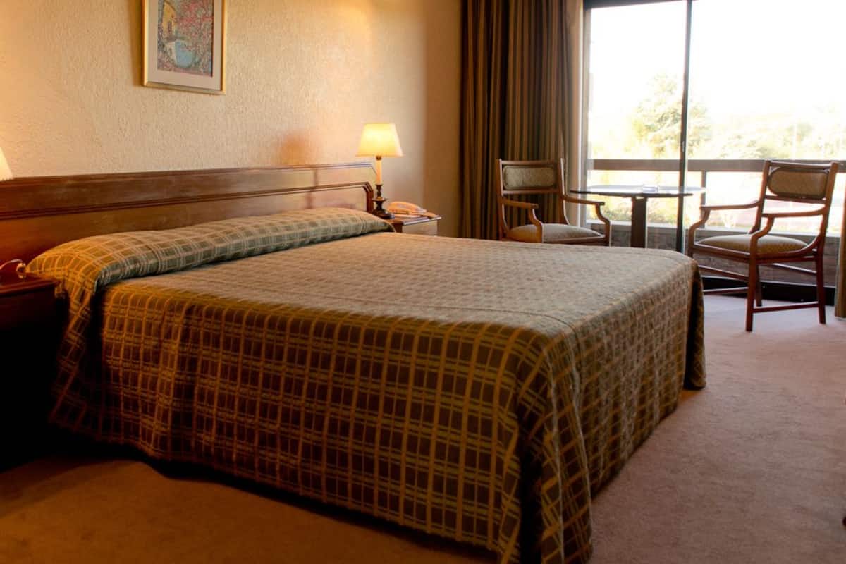 A hotel room with a bed, two bedside lamps, a chair, a small table, and a view of the outdoors.