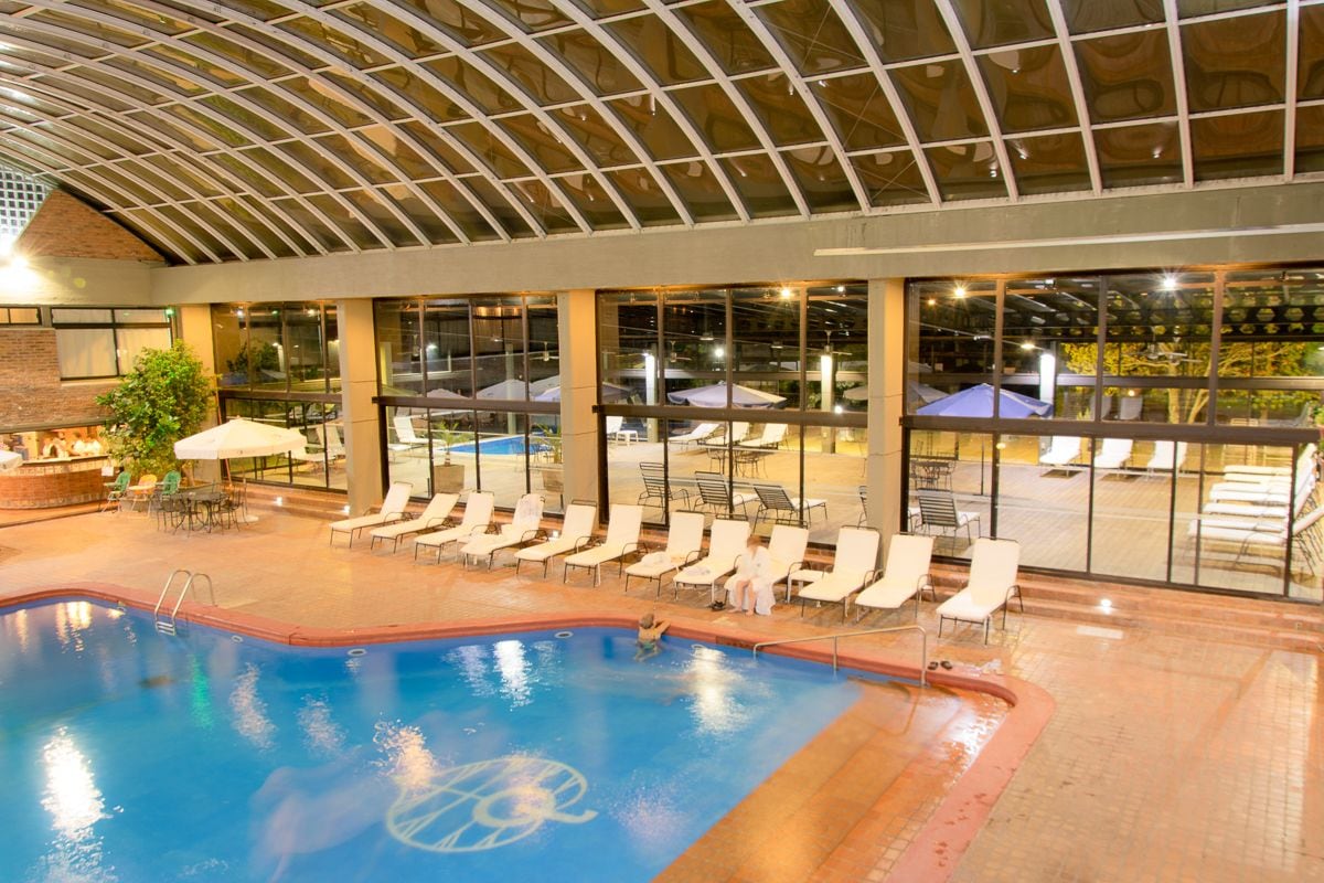 Una piscina interior con sillas de descanso y sombrillas, ventanas amplias y una pared de ladrillo.