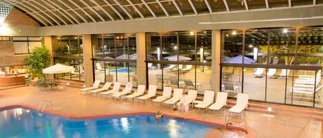 Una piscina interior con sillas de descanso y sombrillas, ventanas amplias y una pared de ladrillo.