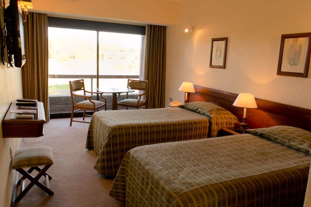 A hotel room with two beds, a desk with a chair, a small table, and a window with curtains.