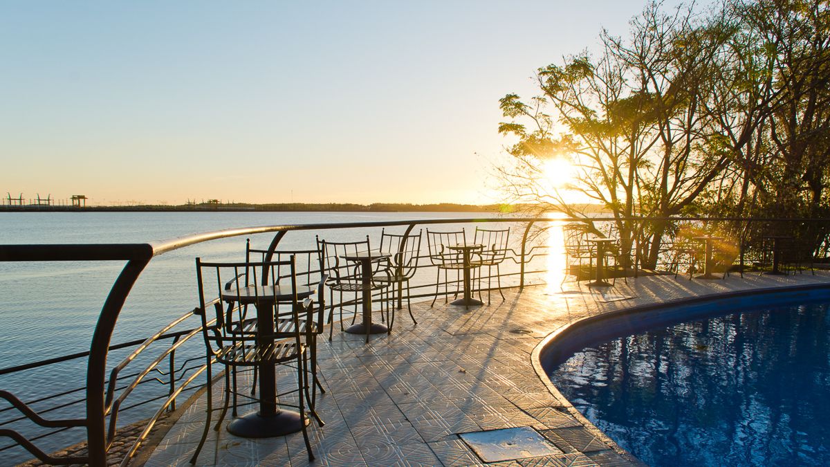 A poolside area with metal chairs and tables, a pool, and a view of the water and horizon during sunset.