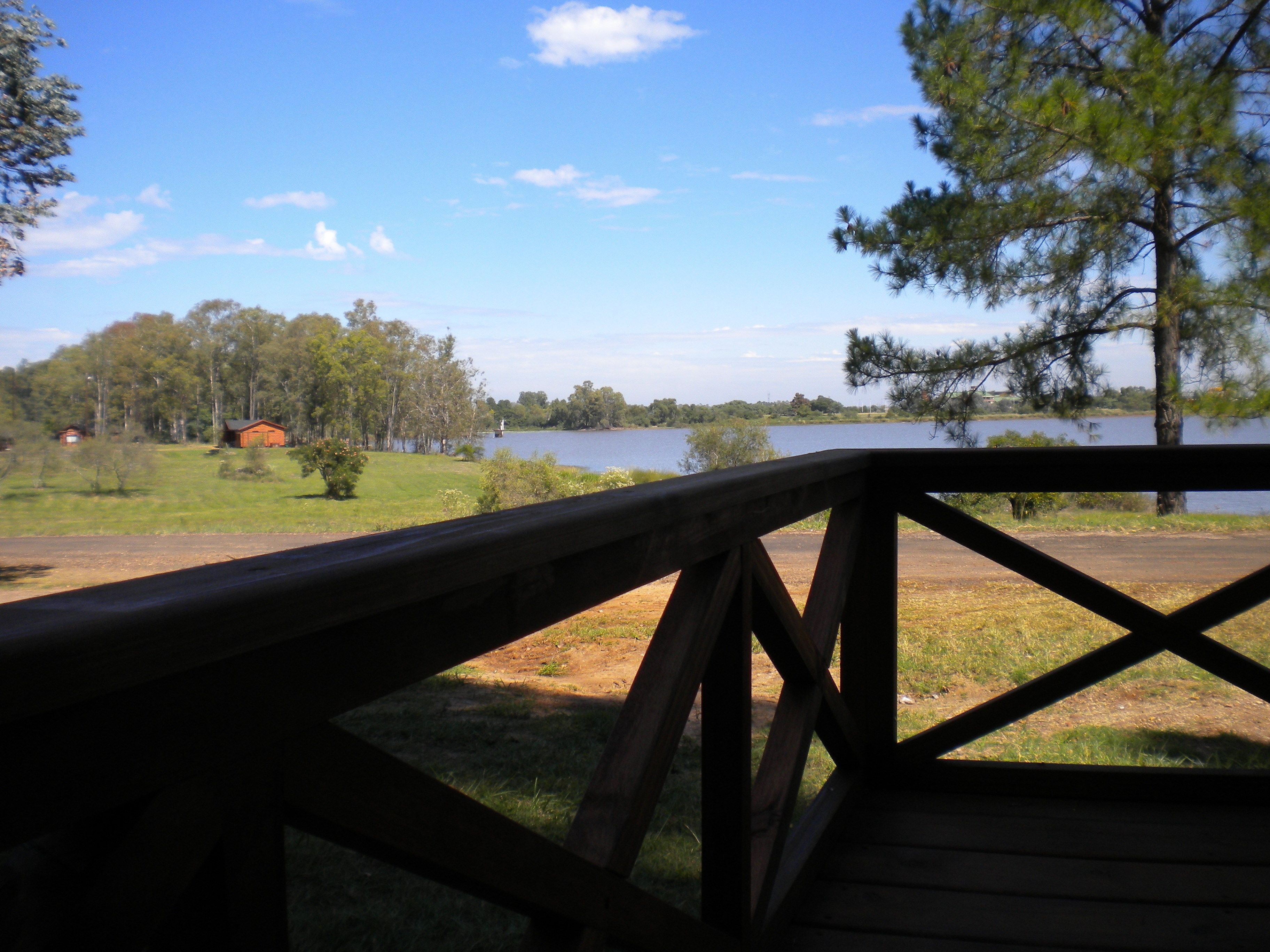 Um deck de madeira com vista para um lago, árvores e uma cabana distante.