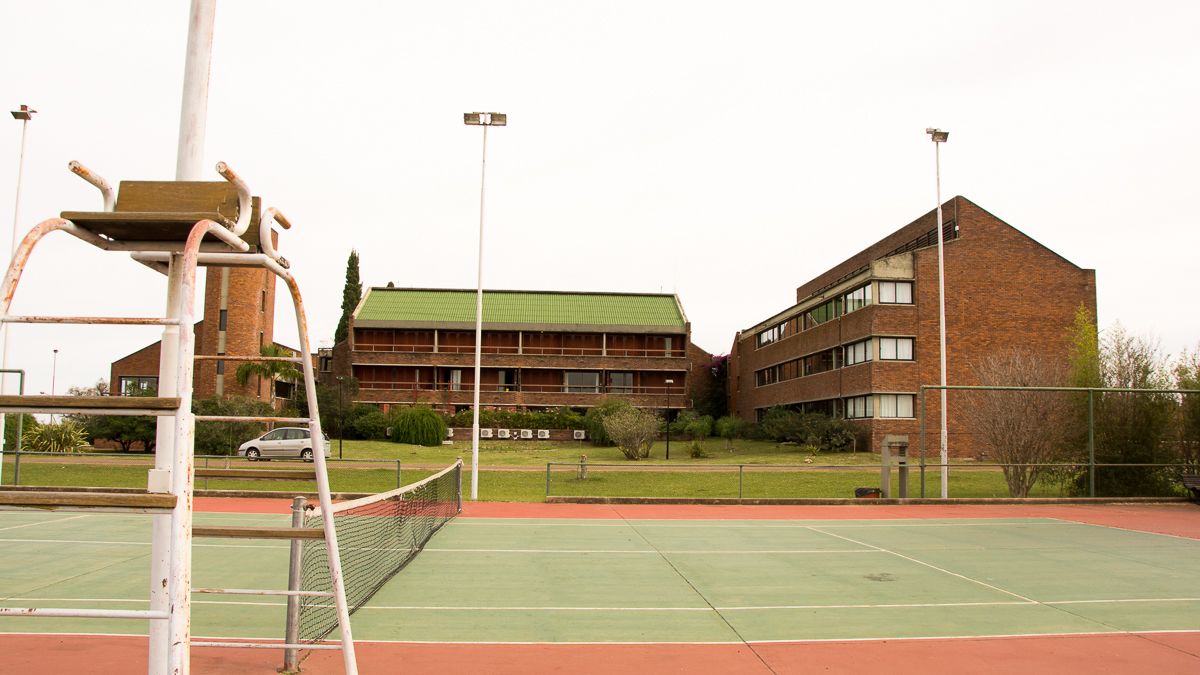 Una cancha de tenis con red, rodeada por una cerca, y un edificio con techo verde al fondo.