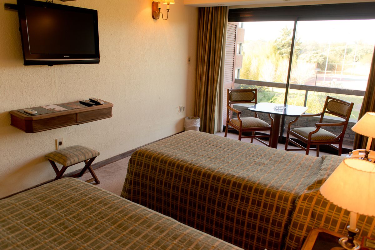 Habitación de hotel con dos camas, televisión, una mesita con sillas y ventana con cortinas.