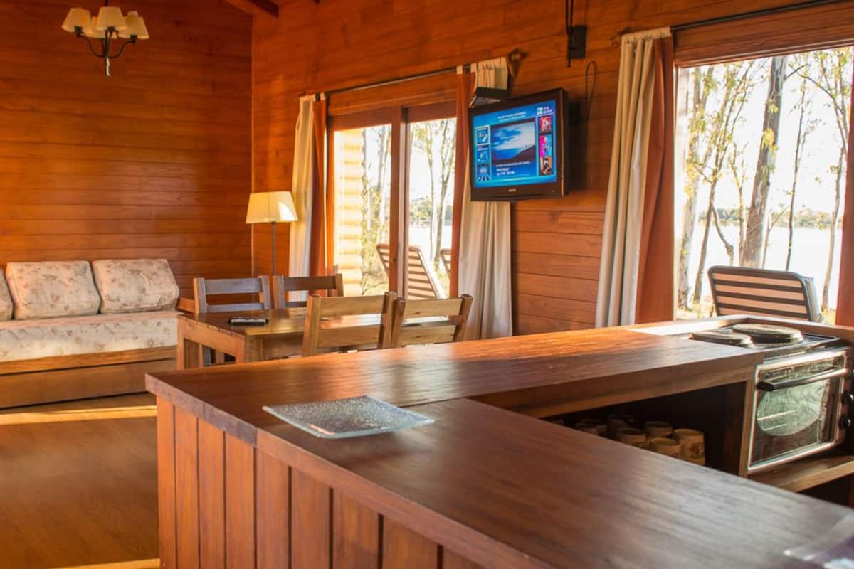 A wooden cabin interior with a sofa, dining table, chairs, a television, and a kitchen area with a stove.