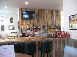 Zona de bar con pared de madera, televisor de pantalla plana y varias botellas. Se observan sillas y una carta.