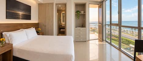A bedroom with a large bed, a nightstand, a wardrobe, a bathroom with a shower, and a balcony with a view of the ocean.