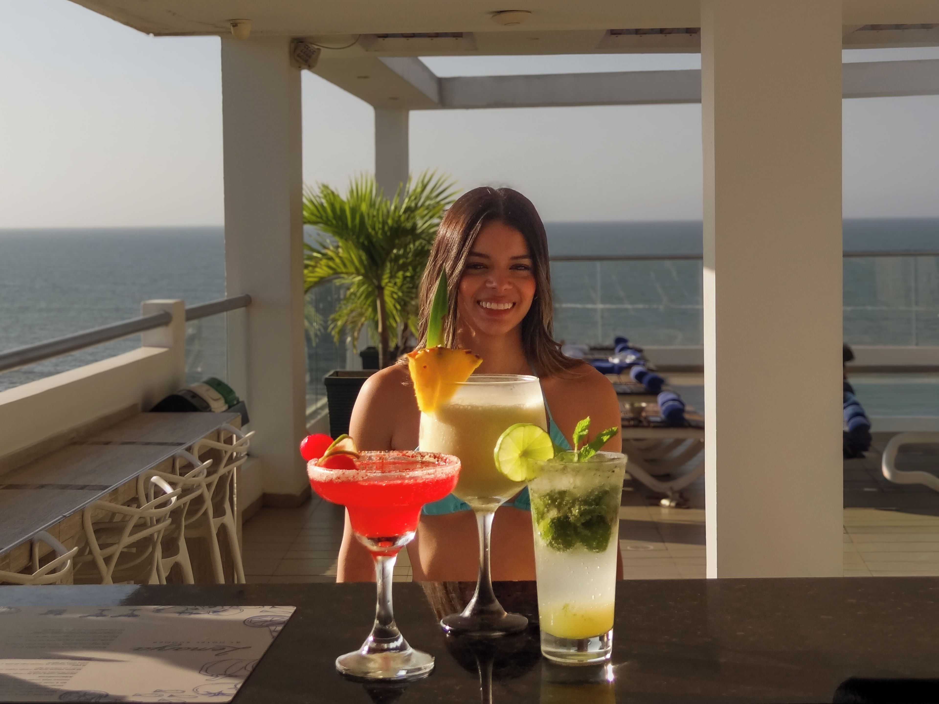 Una persona sentada en una mesa con dos bebidas, vista al mar y un balcón con sillas.