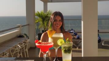 Una persona sentada en una mesa con dos bebidas, vista al mar y un balcón con sillas.