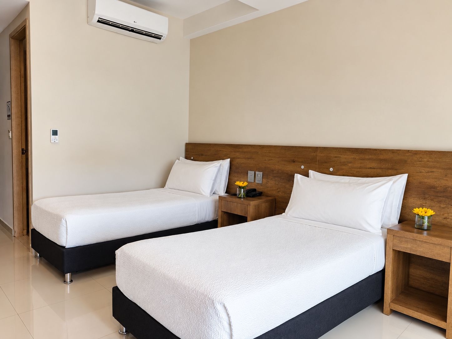 Habitación de hotel con dos camas, cabecera de madera, mesitas de noche con lámparas y aire acondicionado.
