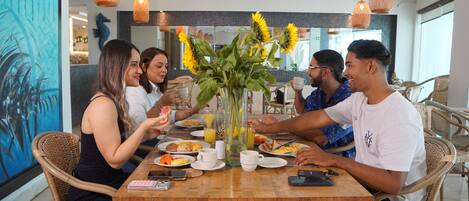 A group of people dining at a wooden table with a vase of sunflowers, surrounded by wicker chairs and orange pendant lights.